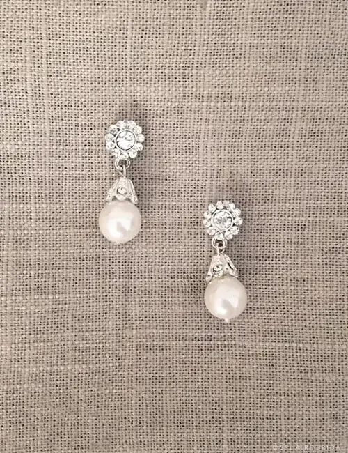 A pair of elegant earrings featuring a round crystal at the top and a pearly drop below, displayed on a textured beige background.