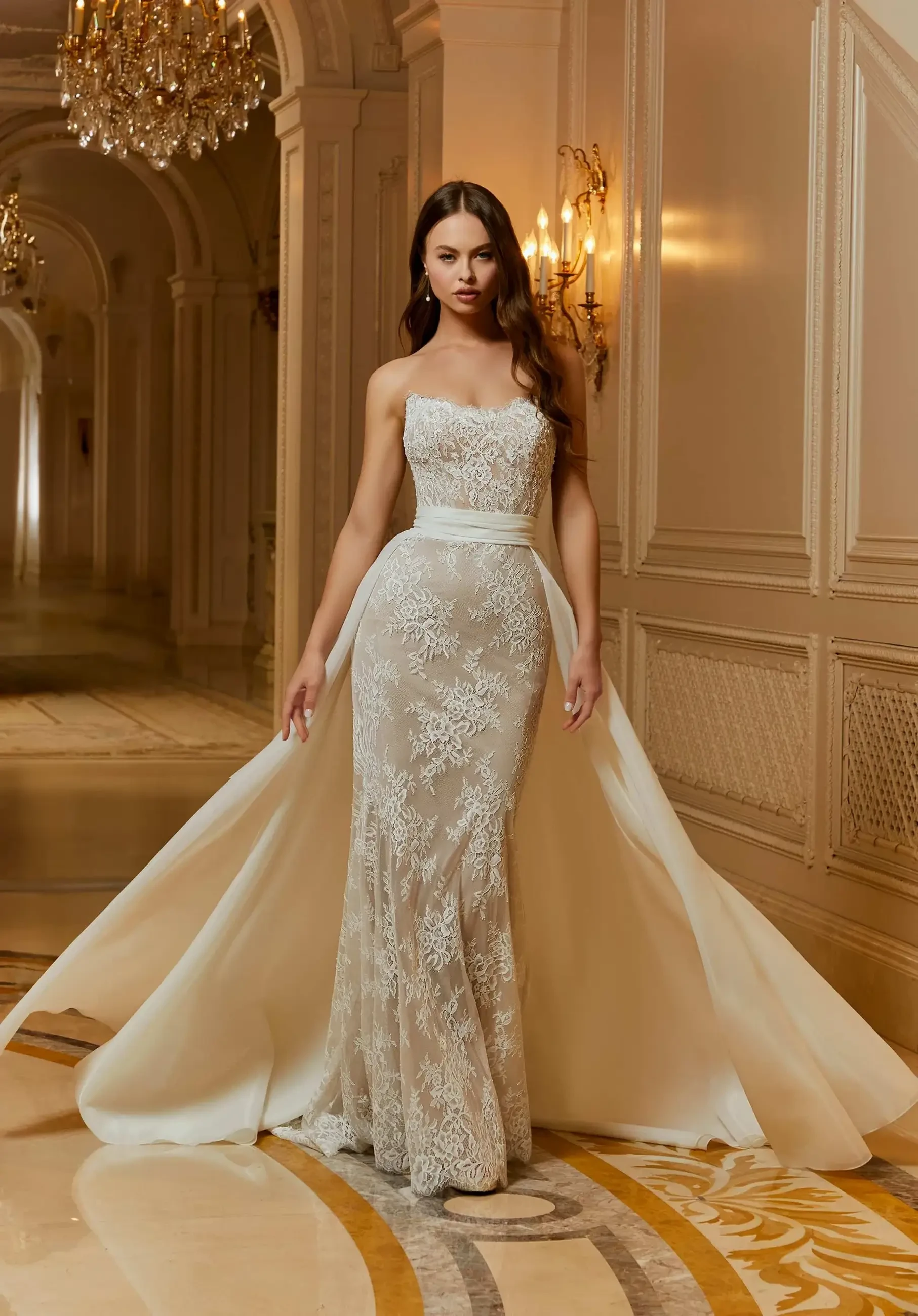 A model wearing a strapless, intricately designed lace gown with a flowing train, standing in an elegant hallway adorned with chandeliers.