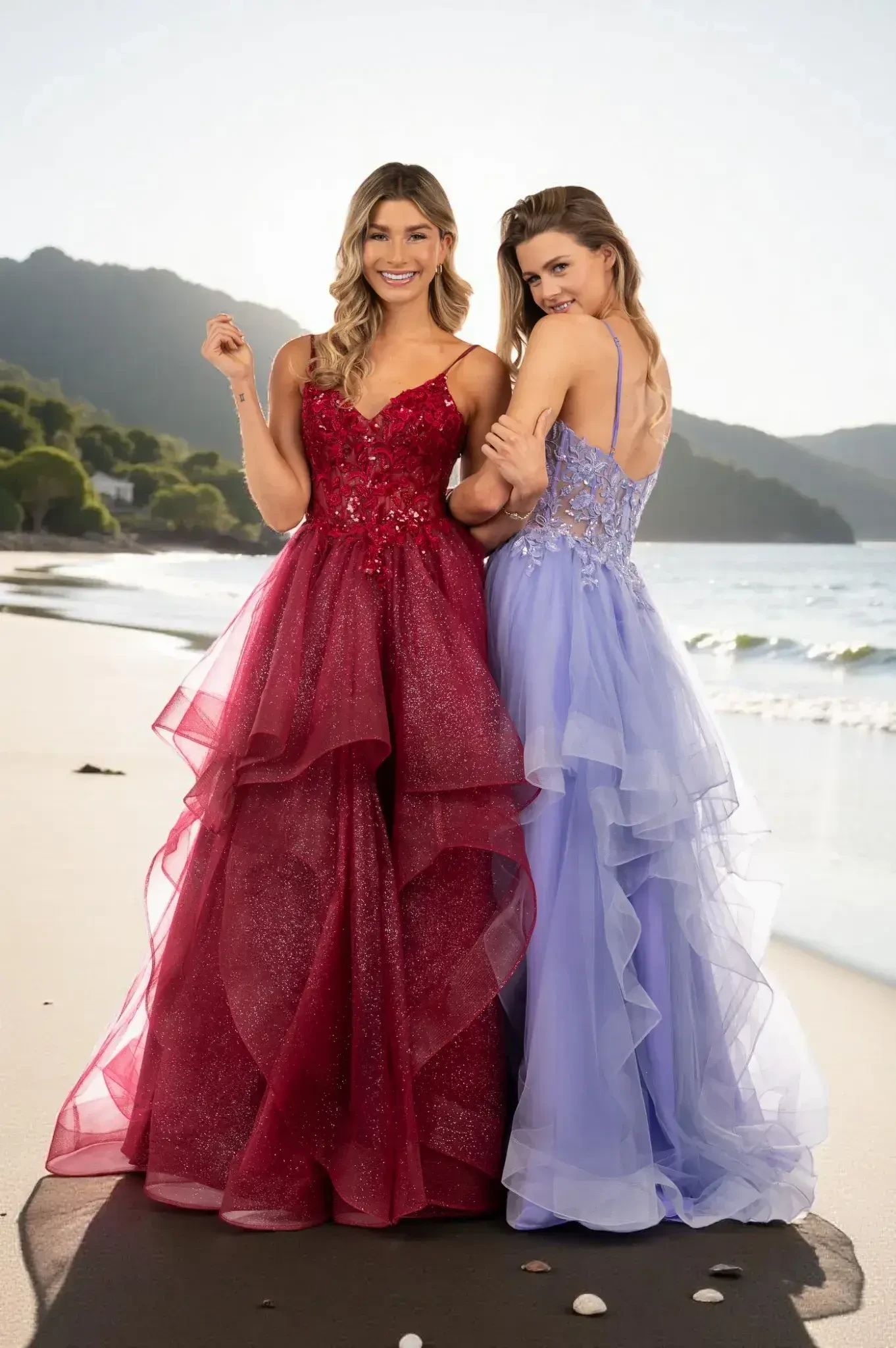 Two women in elegant, flowing dresses stand on a sunlit beach. One wears a glittery red gown, the other a lavender one, both smiling warmly.