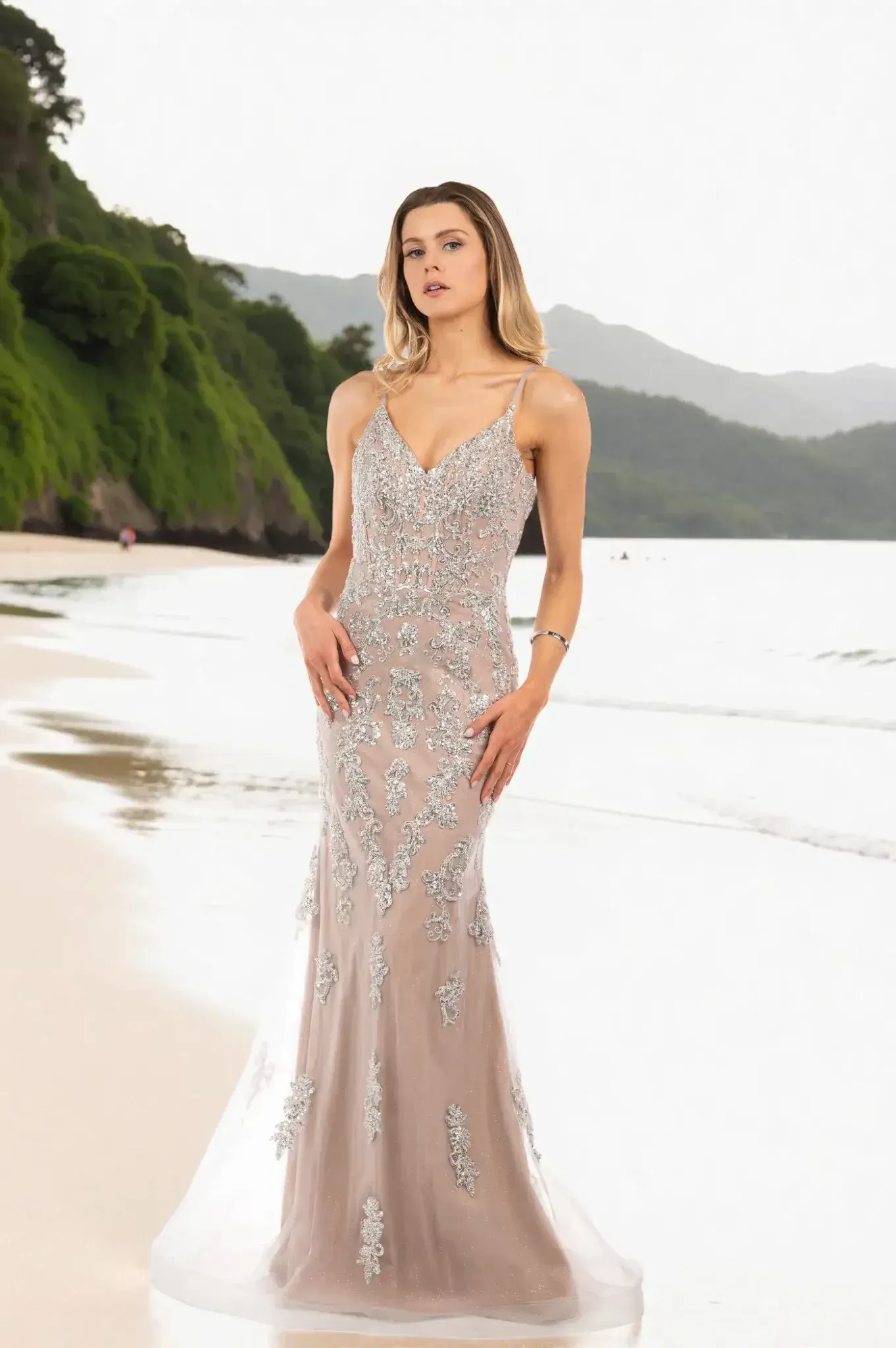 Woman in a silver, embellished gown stands on a serene beach, surrounded by lush green cliffs and gentle waves, conveying elegance and tranquility.