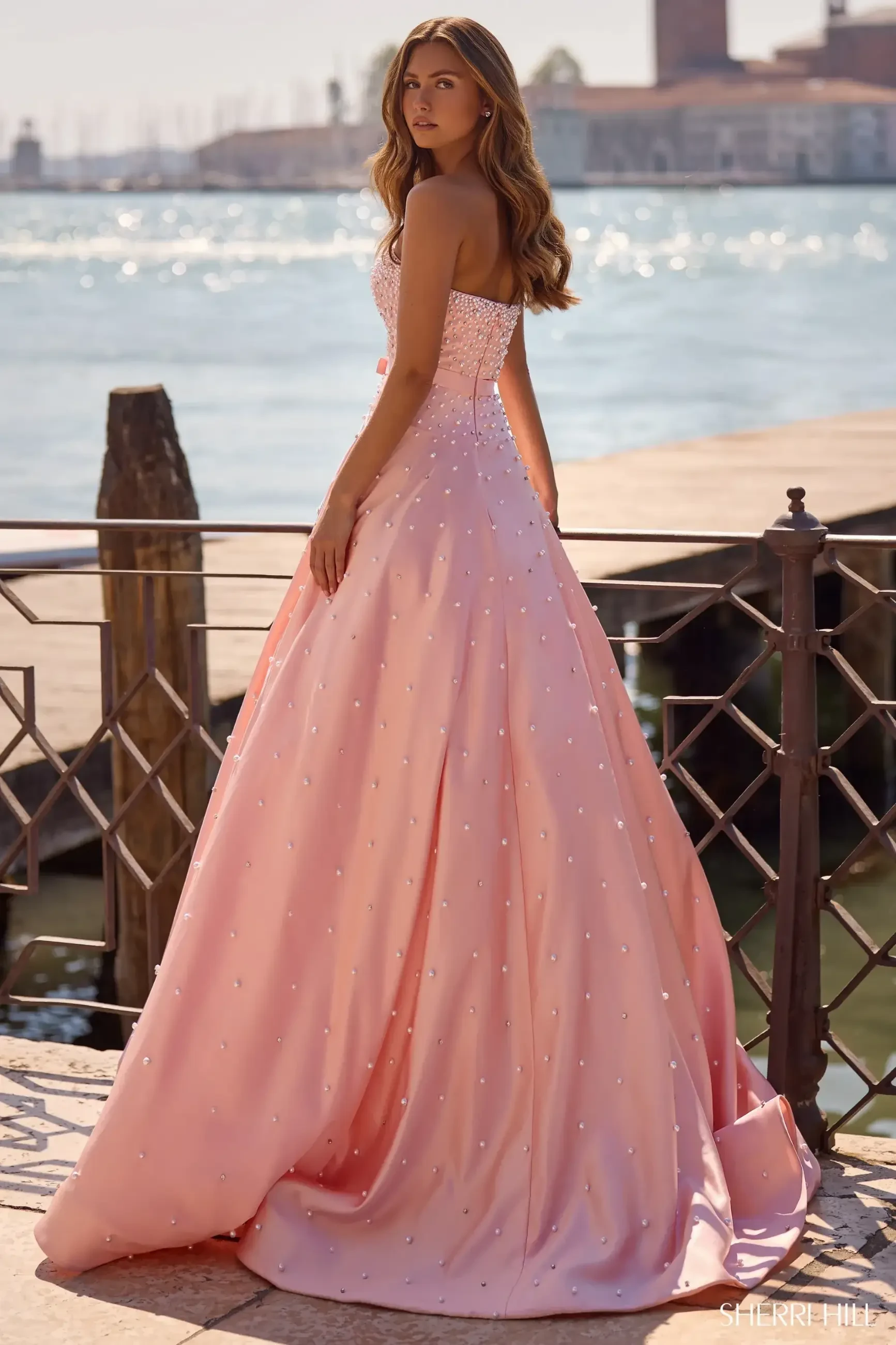 Woman in a strapless pink gown with pearl embellishments stands by a waterfront railing, gazing back. Sunlight sparkles on the water, creating a serene, elegant scene.