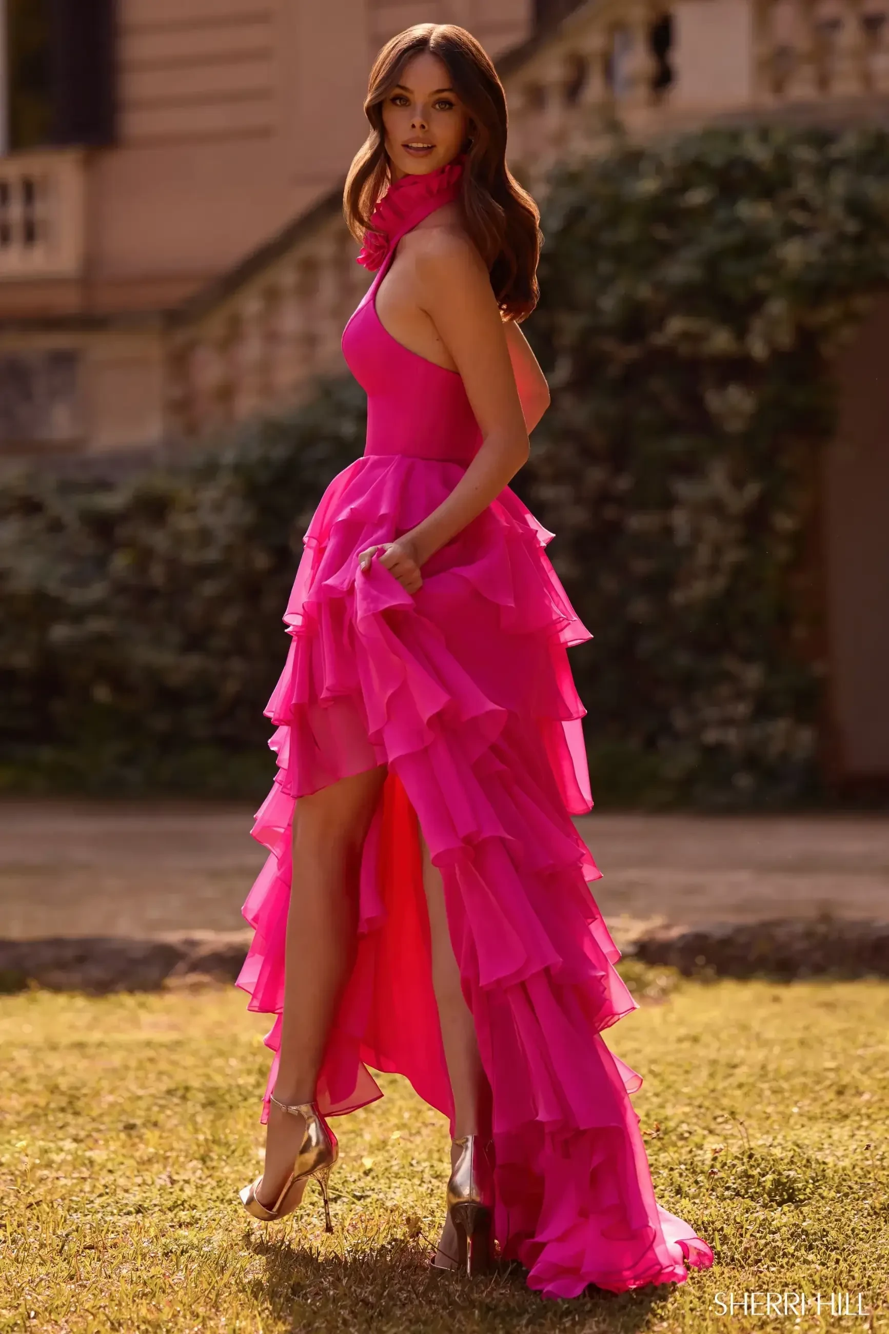 A woman stands outdoors in a vibrant pink, ruffled dress with a high-low hem. She has long brown hair and wears gold heels, exuding elegance and confidence.