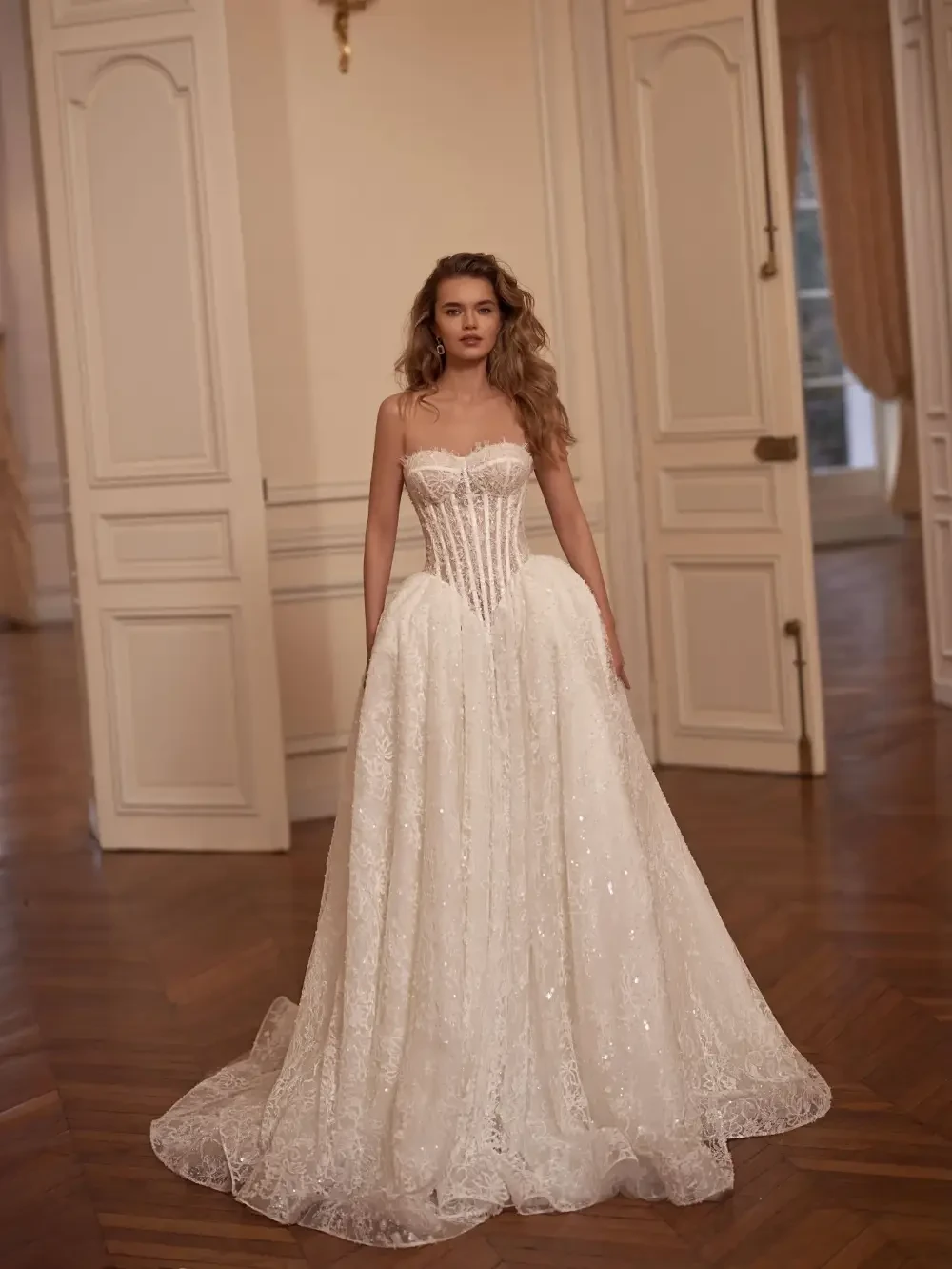 A woman wearing a beautiful white wedding dress with intricate detailing, standing in a grand room with elegant decor.