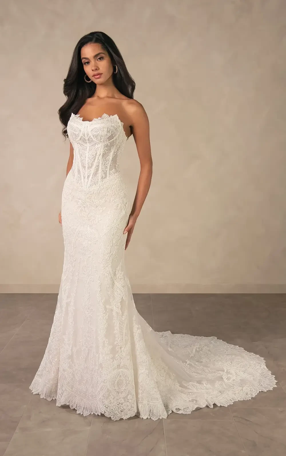 A woman wearing a fitted white wedding dress with a lace design and a train, standing against a neutral background.