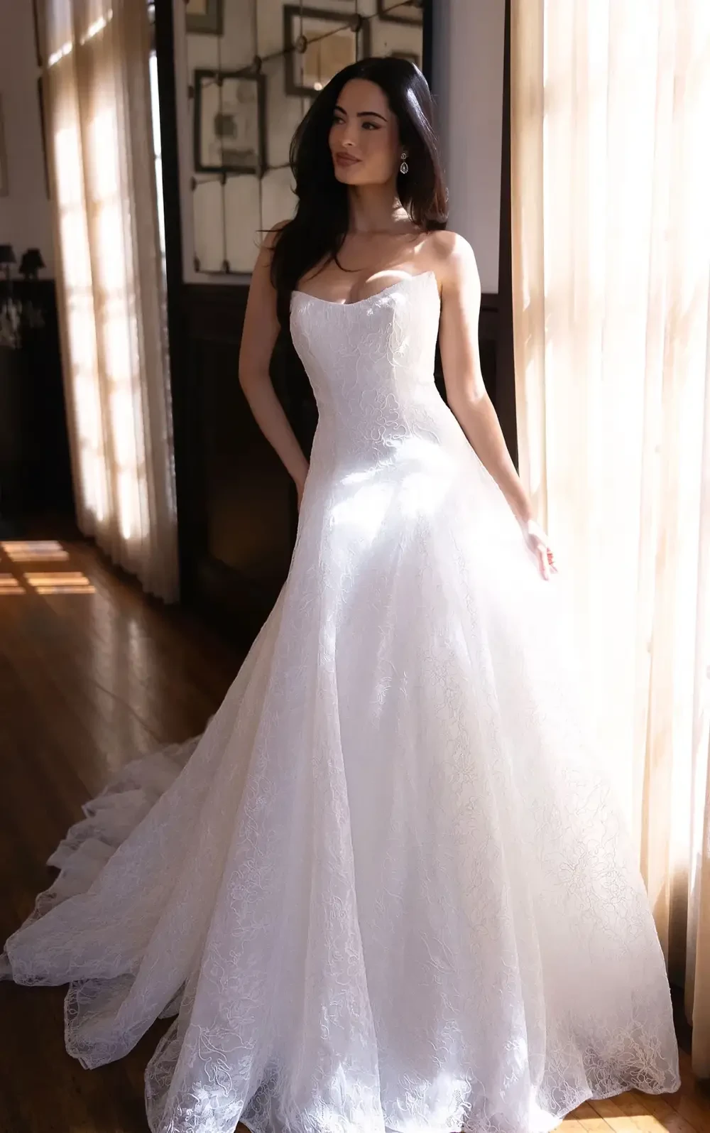 A woman in a beautiful white wedding gown standing in a well-lit room with sheer curtains.