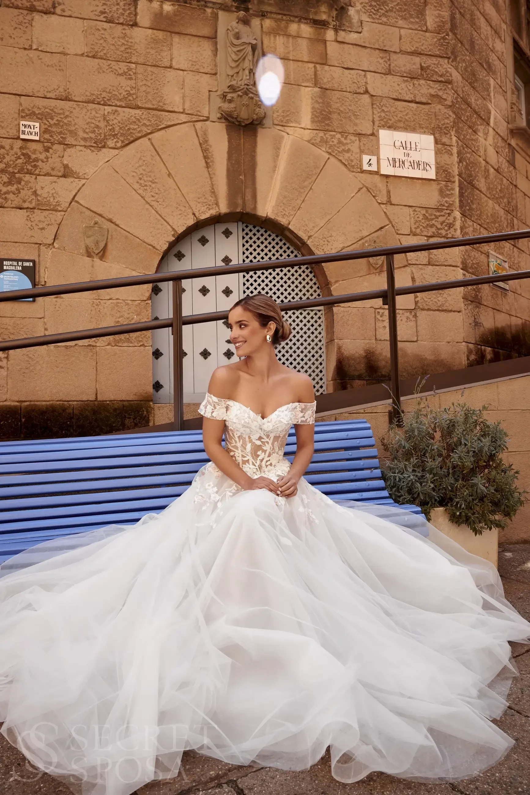 A woman in a white wedding gown with off-the-shoulder sleeves sitting on a blue bench in front of a stone building.