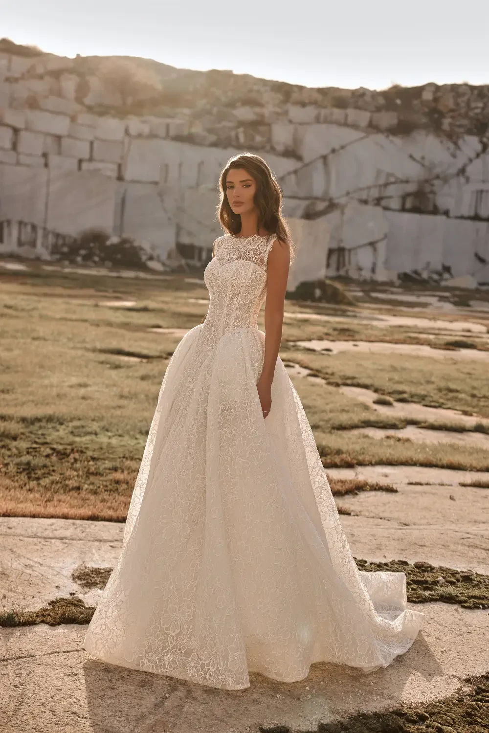 A woman wearing a beautiful, flowing white wedding dress stands in a rocky outdoor setting, with a serene expression and soft sunlight illuminating the scene.