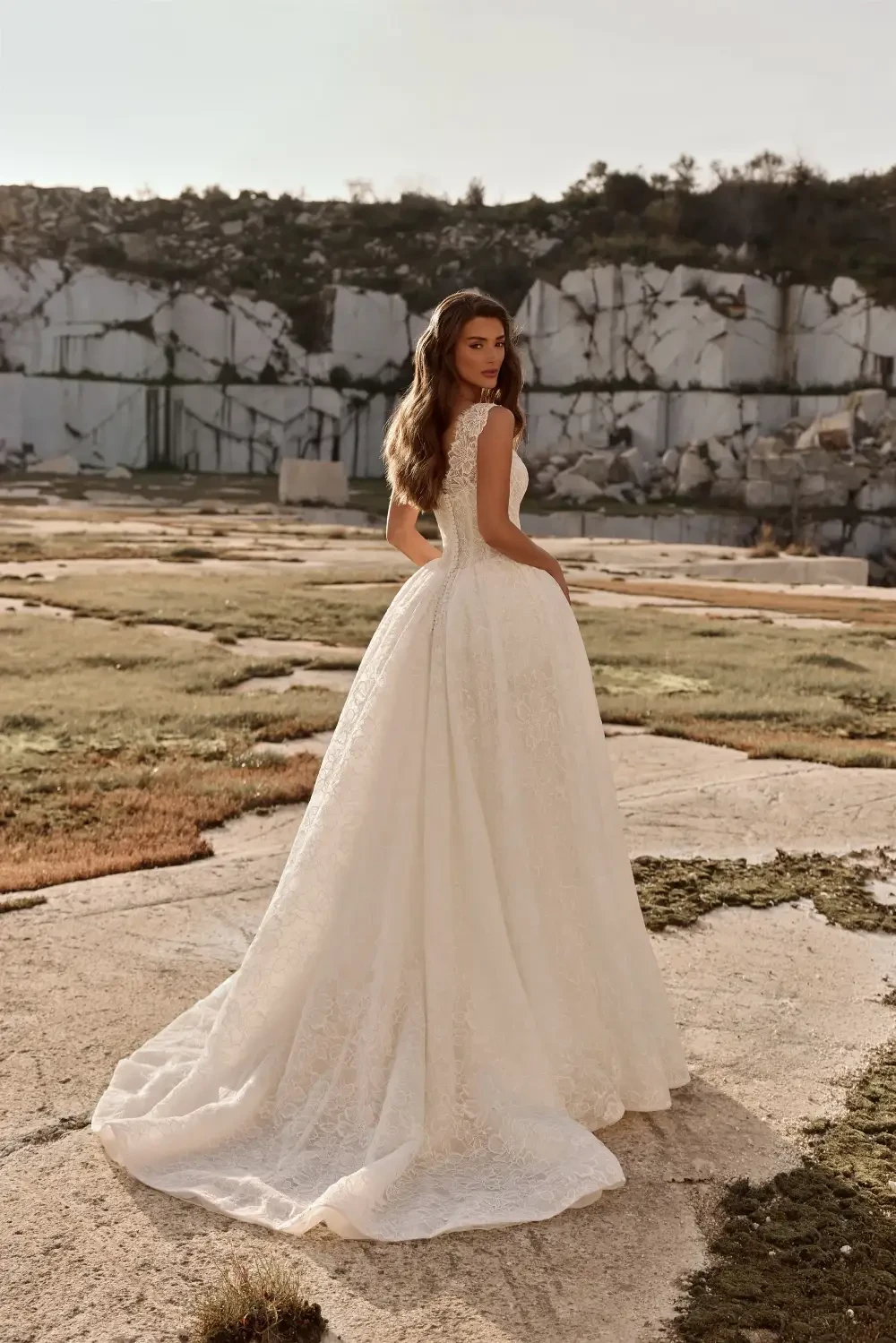 A woman in a beautiful, intricate white wedding gown posing in a scenic outdoor location with rocky terrain.